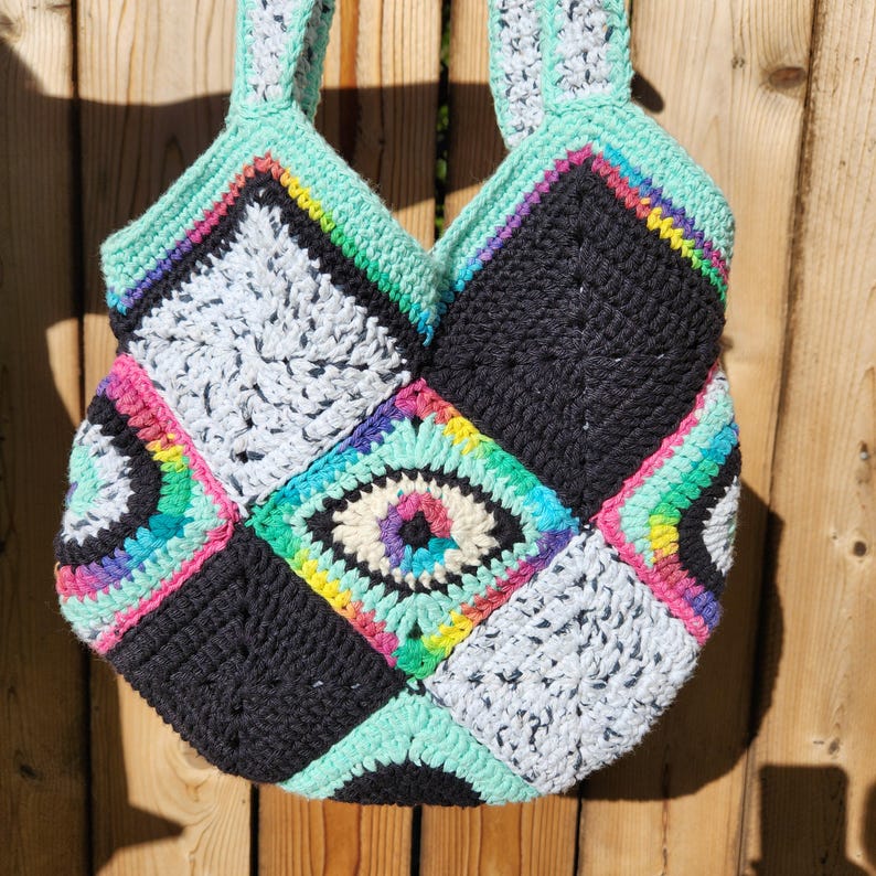 May include: A crocheted tote bag with a patchwork design. The bag is made of black, white, and turquoise yarn with colorful accents. The center of the bag features a large eye design.
