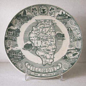 May include: A decorative plate featuring a map of Illinois, with green illustrations of landmarks and the state outline. The plate includes the text "ILLINOIS LAND OF LINCOLN" and is displayed on a clear stand. The plate has a white background.