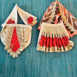 May include: A collection of ornate, miniature fabric garments. The pieces include a cream-colored pouch with red and gold accents, a matching head covering, and a pleated skirt with red and gold trim. An orange and gold sheer shawl completes the set.
