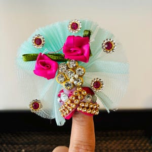 May include: A finger ring with a light blue tulle base, decorated with pink satin roses, gold and white rhinestones, and pink gems.