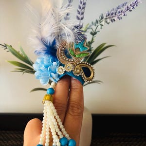 May include: A decorative headpiece featuring blue and white feathers, a blue flower, and a gold and jewel-encrusted embellishment. A beaded tassel with turquoise beads and white pearls hangs from the headpiece. The headpiece is held in a hand.