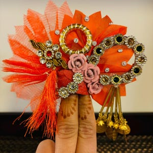 May include: Orange and pink fabric hair ornament with gold and green embellishments, featuring a circular design and a tassel.