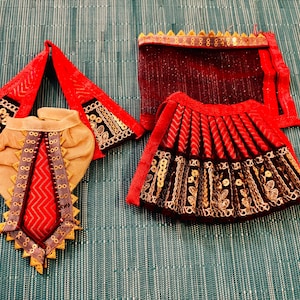 May include: A set of miniature traditional Indian garments. The set includes a red and black skirt, a matching head covering, and a beige and red top. The garments are decorated with gold and silver detailing.