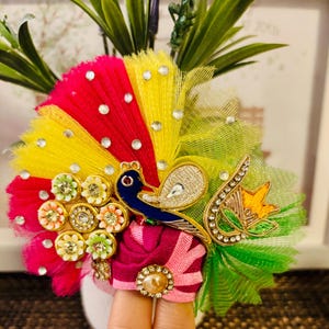 May include: A vibrant, handcrafted peacock-themed accessory. It features a fan-shaped design with red, yellow, and green accents, adorned with rhinestones. A blue peacock figure is centered on a pink and maroon base, embellished with gold trim.