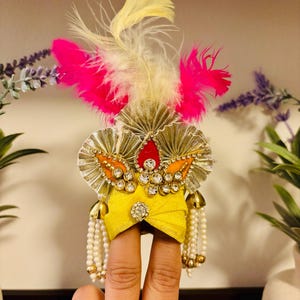May include: A yellow turban-style headpiece with gold embellishments, rhinestones, and a red jewel. It features white and yellow feathers and pink accents. White beaded strands hang from the sides. The headpiece is held up by fingers.