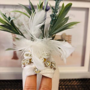 May include: A small, white vase with faux lavender and greenery, accented with white feathers and rhinestone embellishments. The vase is wrapped with a white fabric and adorned with sparkling jewels.