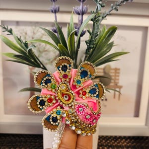 May include: A pink, ornate floral brooch with gold, blue, and brown accents. The brooch features a paisley design, colorful gemstones, and a pearl strand. The brooch is set against a backdrop of lavender and greenery.