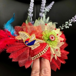 May include: A vibrant, handcrafted headpiece featuring a red, layered fabric base adorned with a gold-embroidered bird, sparkling rhinestones, and colorful feathers. The design incorporates green and yellow floral accents and lavender sprigs.