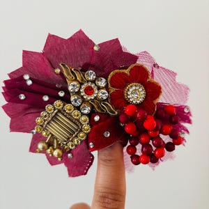 May include: A decorative burgundy and red floral hair accessory. The accessory features a cluster of red berries, a red flower, and a square gold-toned embellishment with gold beads. Clear and red rhinestones are scattered throughout.