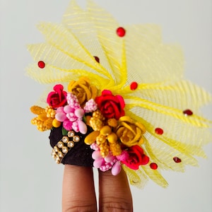 May include: A decorative headpiece featuring a black base adorned with a cluster of colorful flowers in shades of red, pink, and yellow. A yellow mesh fan and red rhinestones add to the design. The headpiece is embellished with gold-colored rhinestones.
