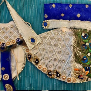 May include: An ornate, cream-colored garment with intricate embellishments, including gold and silver accents, blue jewels, and peacock feathers. The design features a diamond pattern and paisley details. A blue sash and head covering are also included.