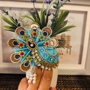 Op de afbeelding: Een sierlijk, turquoisekleurig hoofddeksel met gouden en juwelen accenten. Het hoofddeksel heeft een pauwenveerontwerp met parels die aan de onderkant hangen. Het hoofddeksel staat voor een vaas met lavendel en groene bladeren.