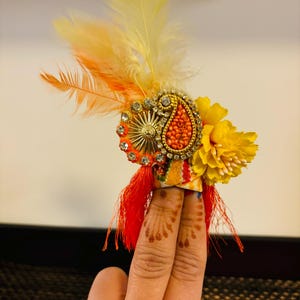 May include: A colorful finger ring with a paisley design, yellow flower, and red tassels. The ring is decorated with rhinestones and beads. The ring is worn on two fingers.
