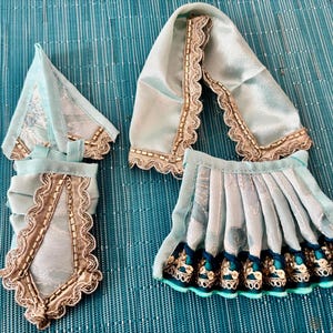 May include: A set of doll clothing in shades of light blue, featuring intricate embroidery and beadwork. The set includes a top, a shawl, and a pleated skirt, all with decorative trim. The details suggest a traditional or cultural design.