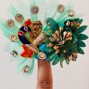 May include: A decorative headpiece featuring a peacock design with blue, red, and gold accents. The headpiece includes a green mesh base, sparkling rhinestones, and a teal and gold floral arrangement. The design is intricate and ornate.