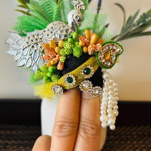 May include: A decorative floral arrangement featuring a silver peacock, green mesh, and various colorful beads. The arrangement includes a black and yellow band adorned with green and clear gemstones, and a strand of white pearls.