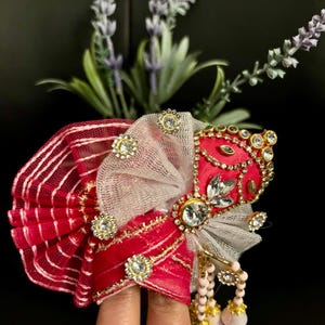 May include: A decorative headpiece featuring a vibrant red and white striped fabric, accented with gold trim and sparkling rhinestones. The headpiece has a central red section adorned with more rhinestones and a delicate gray mesh overlay. Pink beads dangle from the bottom.