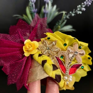 May include: A vibrant, handmade hair accessory featuring a combination of textures and colors. The design includes a yellow fabric base adorned with gold embroidery, red accents, and faux pearls. A burgundy mesh bow and a yellow rose add to the decorative flair.