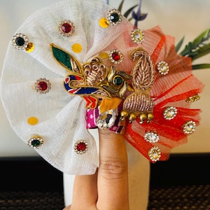 May include: A decorative headpiece with white and red fabric, adorned with colorful jewels and gold embellishments. The design features a peacock motif and is accented with yellow polka dots. The headpiece is held by a finger.
