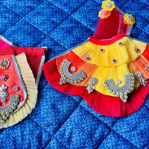 May include: Two ornate, colorful garments with intricate detailing. One is a red and yellow skirt-like piece with silver embellishments. The other is a matching red and pink pouch with silver trim and decorative elements. Both are on a blue patterned surface.
