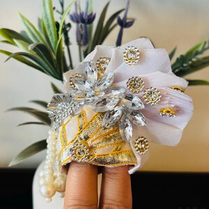 May include: A white and gold fabric headpiece adorned with clear and gold-toned rhinestones. The headpiece features a floral design with a pearl strand and is set against a backdrop of lavender and greenery.