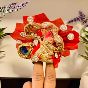 May include: A decorative, ornate red and gold finger ring with a peacock feather accent. The ring features intricate beadwork, sparkling rhinestones, and a layered design. The ring is held up by two fingers.