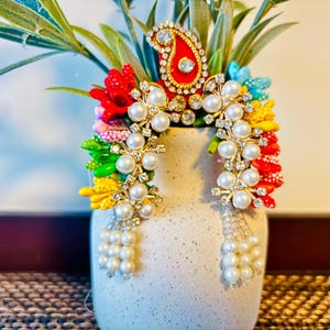 May include: A decorative headpiece with a red and gold paisley design, adorned with clear rhinestones and pearls. The headpiece is embellished with colorful floral accents and pearl dangles. The piece is displayed in a white vase with green foliage.
