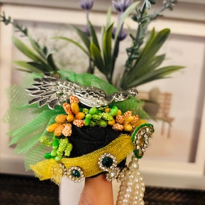May include: A decorative brooch featuring a silver peacock design, green and yellow accents, and faux pearls. The brooch has a black base with green and orange floral embellishments and is adorned with green gemstones.