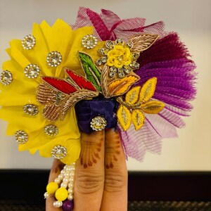 May include: A colorful flower crown with yellow, purple, and green fabric, embellished with rhinestones and gold embroidery. The crown is decorated with a small yellow flower and a pearl and bead tassel.