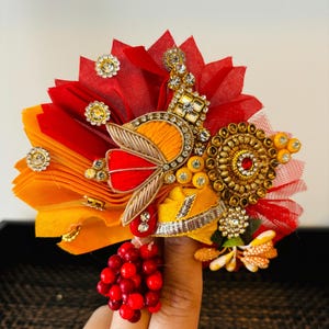 May include: A colorful crown made of fabric, beads, and rhinestones. The crown is decorated with a red, orange, and yellow fabric base, and features a gold and silver design with a red and gold butterfly motif. The crown is accented with red beads and rhinestones.