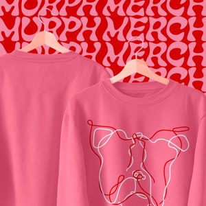 May include: Two pink sweatshirts, one with a white line drawing of a dog's face and the other plain. Both sweatshirts are hanging on hangers against a red background with a repeating pattern of the word "MERCH" in a wavy font.