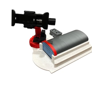 May include: A black phone holder with a red base and a small grey structure with two circular openings. A grey building model with a red trim and blue grid windows is also present. The objects are on a white surface.