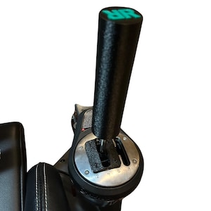 May include: Black gear shift lever with a textured grip and a silver base. The top of the lever has the letters "MR" in green. The base has gear number markings. The lever is part of a racing simulator setup.