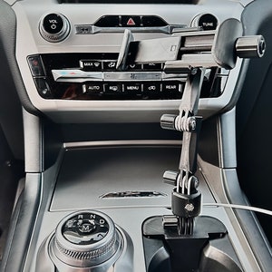 Op de afbeelding: Een zwarte, verstelbare telefoonhouder geïnstalleerd in de middenconsole van een auto. De houder is bevestigd aan de bekerhouder en heeft meerdere gewrichten en verstelknoppen. Het interieur van de auto heeft een zilver en zwart kleurenschema.