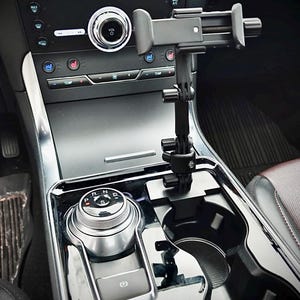 May include: A black car phone holder mounted in the centre console. The holder is attached to the cup holder area and extends upwards. The car's dashboard and gear shift are visible in the background.