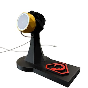 Op de afbeelding: Een zwarte 3D-geprinte oplaadstandaard met een gouden en witte slimme speaker erop. De standaard heeft een rood schorpioenlogo op de basis.