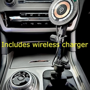 以下が含まれることがあります： 車のシフトレバーに取り付けられた黒いワイヤレス携帯電話充電器。充電器は、銅色のコイルが見える円形の充電パッドを持っています。画像には「Includes wireless charger」というテキストが表示されています。