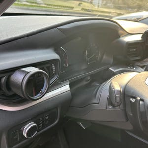 以下が含まれることがあります： 車のインテリアのクローズアップ。黒いダッシュボードに、左側に丸いゲージが取り付けられている。ゲージは白い文字盤で、黒い針が付いている。背景にはステアリングホイールが見える。