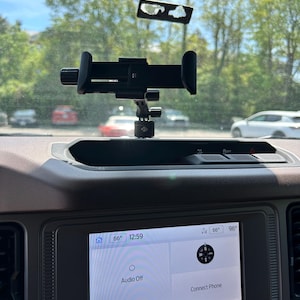 Può includere: Un supporto nero per telefono da auto montato sul cruscotto di un'auto. Il supporto è regolabile e ha un alloggiamento per fissare un telefono. Lo schermo di infotainment dell'auto mostra l'ora, la temperatura e le impostazioni audio.