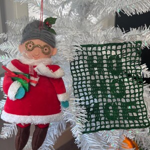 May include: A felt Christmas ornament of a woman wearing glasses and a red dress holding a wrapped gift. The ornament is hanging on a white artificial Christmas tree with a green crocheted square ornament hanging next to it.