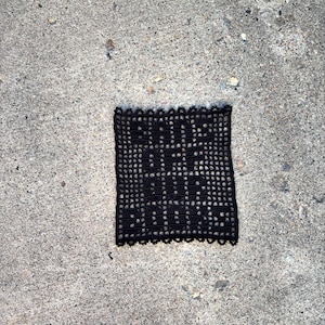 Könnte beinhalten: Schwarze Häkeldeckchen mit einem gewellten Rand. Das Deckchen hat ein sich wiederholendes Muster aus kleinen Kreisen und Quadraten.