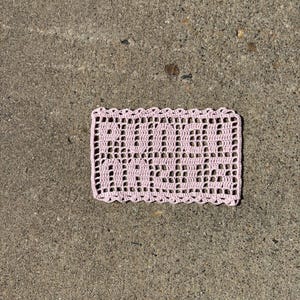 Puede incluir: Una pieza rectangular de ganchillo rosa claro con las palabras "RUDE BABES" tejidas en el diseño. La pieza de ganchillo tiene un borde festoneado y está sobre una superficie de hormigón.