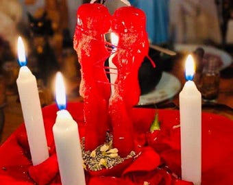 May include: A spiritual arrangement featuring two large red candles intertwined, with a smaller white candle on each side. The candles are lit, and surrounded by red rose petals and herbs on a red surface. Statues are visible in the background.