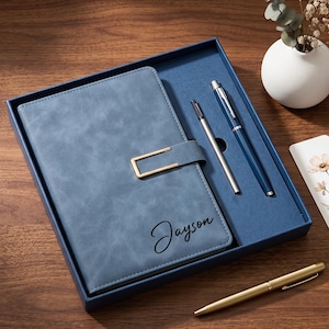 Op de afbeelding: Een blauw notitieboek met een goudkleurige rechthoekige sluiting en de naam Jayson in zwarte schrijfletters. De set bevat een zilveren, een blauwe en een gouden pen, gepresenteerd in een blauwe doos met een bloemrijke boekenlegger.