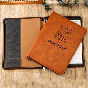 Może przedstawiać: Brązowy skórzany notatnik z imieniem "Layla Brown" i inicjałami "LB" z kwiatowymi akcentami. Notatnik jest otwarty, ukazując papier w linie. Lewa strona notatnika zawiera imię "Alice" i cytat.