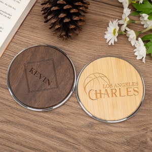 Könnte beinhalten: Zwei runde Holz-Handy-Ladegeräte mit einem silbernen Metallrand. Ein Ladegerät ist braun mit dem Namen "KEVIN" in einem Quadrat eingraviert. Das andere Ladegerät ist hellbraun mit einem Basketball und dem Text "LOS ANGELES CHARLES" eingraviert.