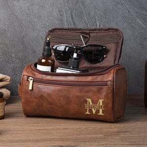 Op de afbeelding: Een bruine leren toilettas met een gouden rits en de initialen "M" en "Mitchell IV" op de voorkant. De tas is open en onthult een zonnebril, een fles en andere toiletartikelen. De tas staat op een houten oppervlak.