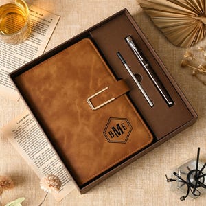 May include: A brown notebook with a gold-colored metal clasp and a black hexagonal monogram featuring the letters BME, presented in a gift box alongside a silver-toned pen and a black ink refill cartridge, arranged on a textured surface.