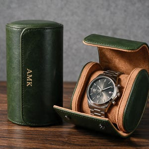 May include: A cylindrical dark green watch case with the letters AMK embossed on the front, paired with an open matching case containing a silver-toned analog wristwatch with a dark grey dial and a metal link bracelet, featuring a tan interior.