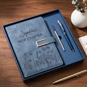 May include: A blue notebook with a gold-tone metal clasp, featuring the text "Sophia's NEW CHAPTER" and a black floral line art design. The set includes two blue pens, one silver pen, and a white bookmark with a floral illustration.